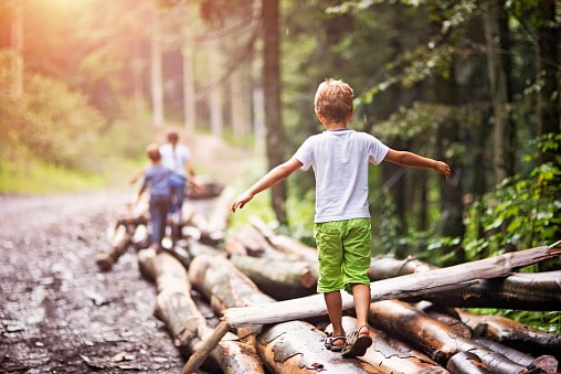 Children Pediatric Balance Disorder Child walking on logs behind parents who are about 50 feet ahead of it. Everyone is in the woods on a gravel logging trail.